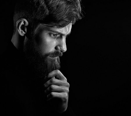 Black and white portrait of puzzled young man touching beard looking down over black background